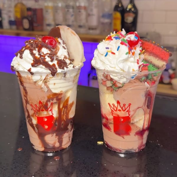 Two BRENDOS milkshakes with whipped cream, chocolate drizzle, sprinkles, and a cherry on a bar.