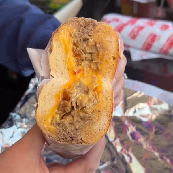 A person holds a BRENDOS cheesesteak sandwich, cut in half, revealing melted cheese and steak.