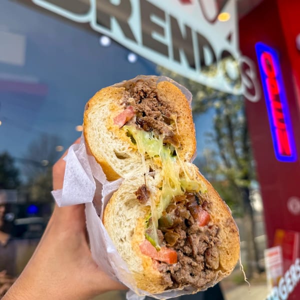 A hand holds a BRENDOS cheesesteak sandwich, cut in half, with melted cheese stretching between the halves.