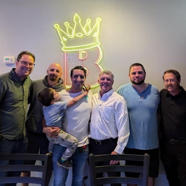 Six men and a young child pose in front of a neon BRENDOS crown logo.