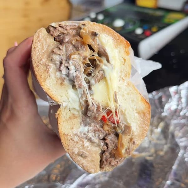 A hand holds a BRENDOS sandwich, cut in half, revealing melted cheese, meat, and red peppers.