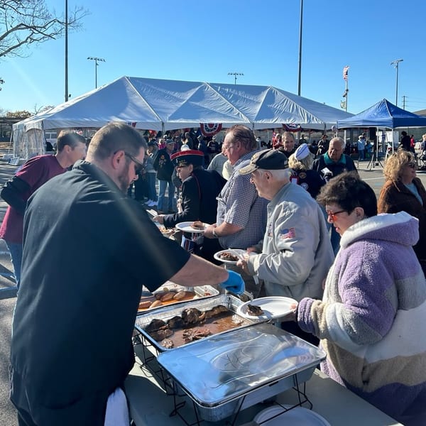 People lining up for food served from chafing dishes under a white tent at an outdoor event.