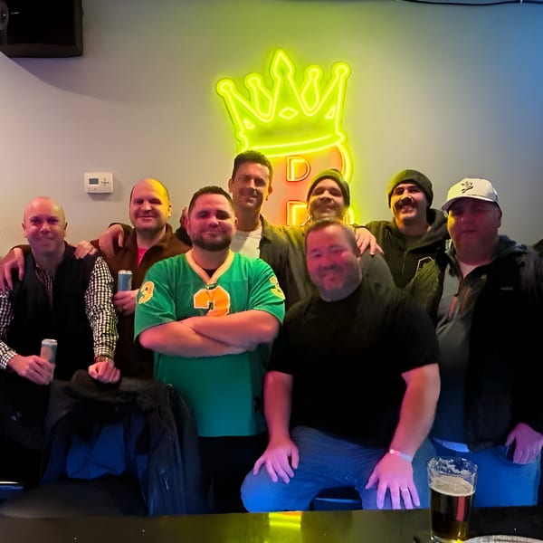 Group of men posing in front of a neon BRENDOS crown logo, holding drinks.
