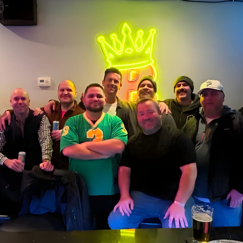 Group of men posing in front of a neon BRENDOS crown logo, holding drinks.