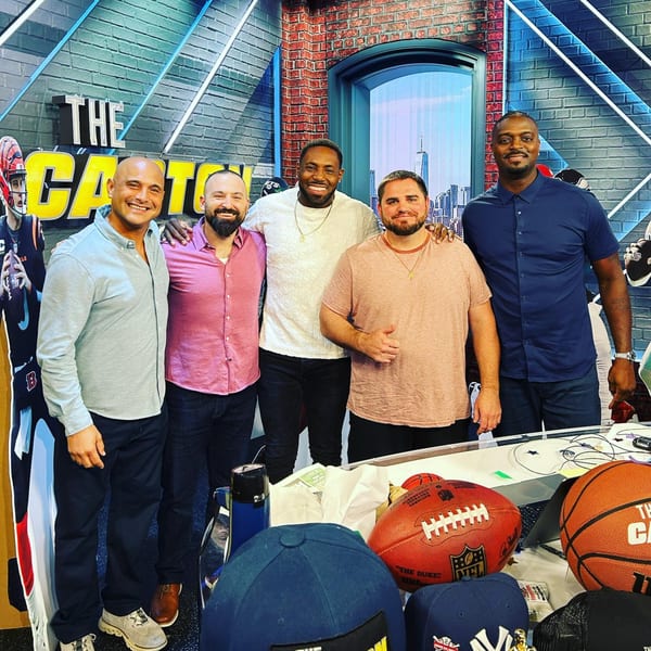 Five men smiling and posing together in a sports-themed studio with sports memorabilia.