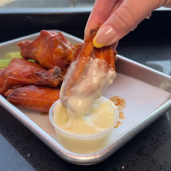 A hand dips a buffalo chicken wing into a small cup of creamy white sauce, with more wings on a tray.