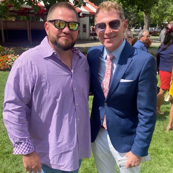 Two men in sunglasses, one in a purple shirt and the other in a blue suit, stand outdoors.