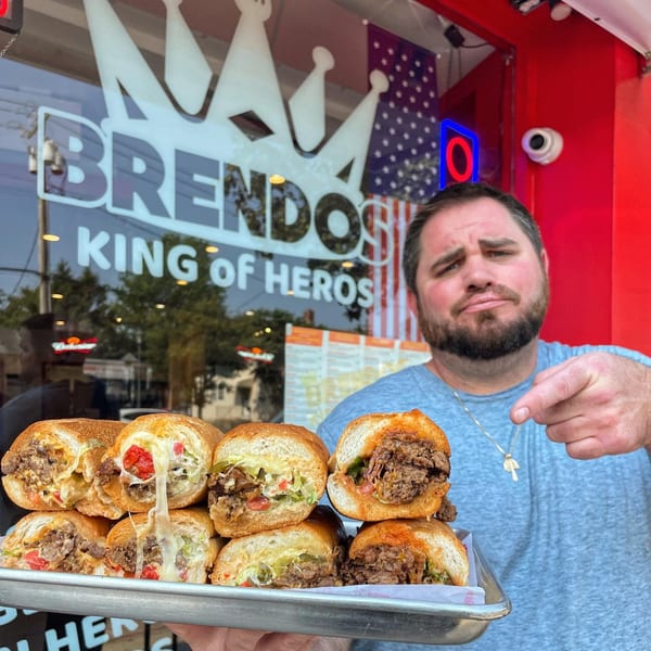 Man holding a tray of BRENDOS cheesesteak sandwiches with melted cheese, pointing at the camera.