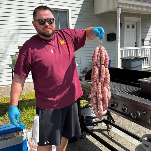 Man in sunglasses and a BRENDOS shirt holding a large string of sausages next to a grill.