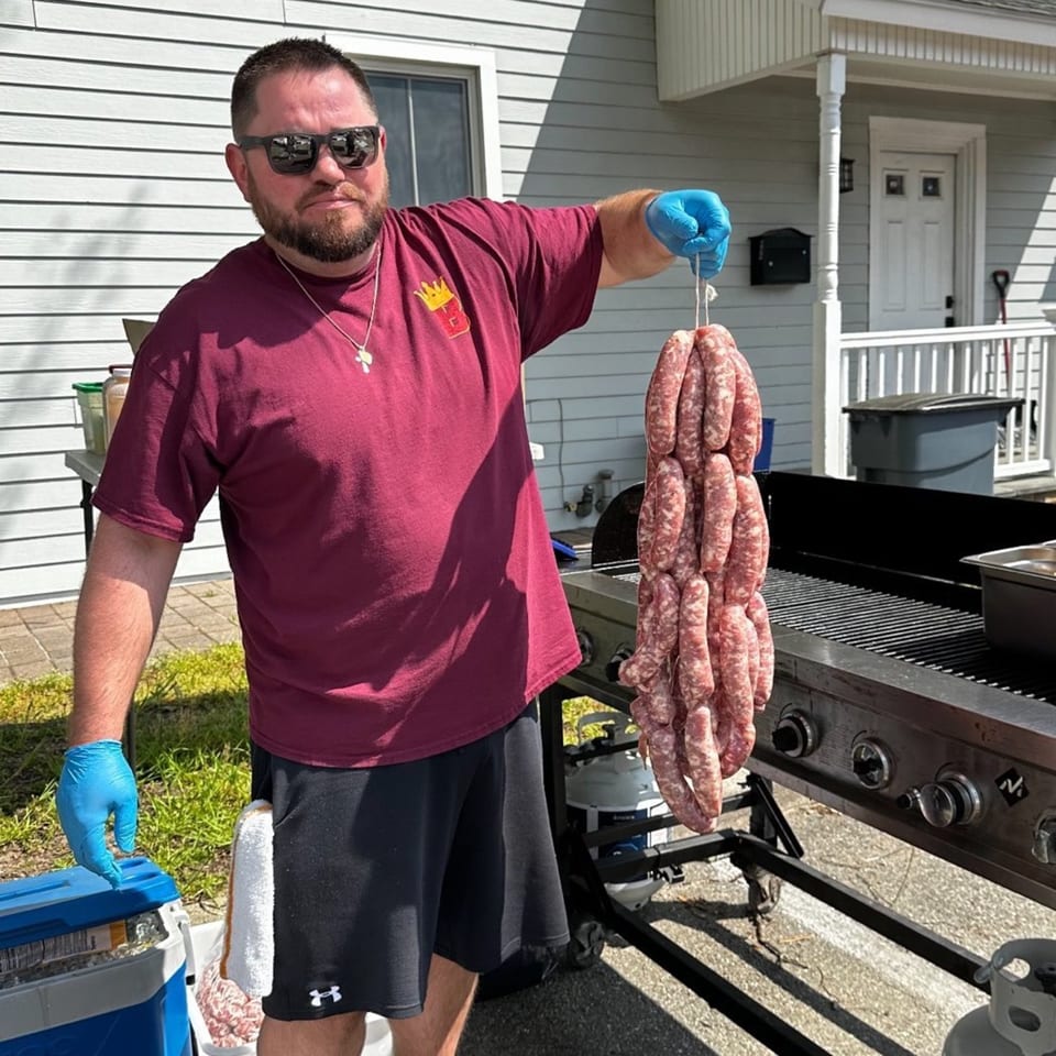 Man in sunglasses and a BRENDOS shirt holding a large string of sausages next to a grill.